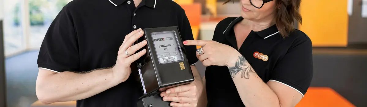 Ein digitaler Stromzähler wird von zwei Personen in schwarzen Hemden gehalten. Eine Person zeigt auf das Display des Zählers. Im Hintergrund sind bunte, verschwommene Formen erkennbar.
