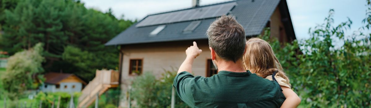 Ein Mann hält ein kleines Mädchen im Arm und zeigt auf ein Haus mit einer Solaranlage in einer grünen Umgebung.