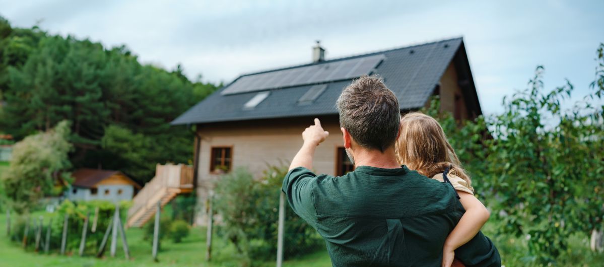 Ein Mann hält ein kleines Mädchen im Arm und zeigt auf ein Haus mit einer Solaranlage in einer grünen Umgebung.