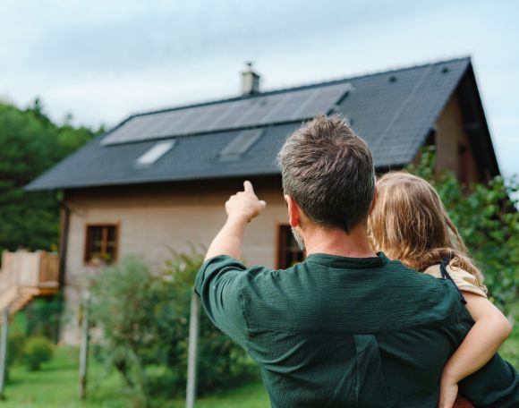 Ein Mann hält ein kleines Mädchen im Arm und zeigt auf ein Haus mit einer Solaranlage in einer grünen Umgebung.