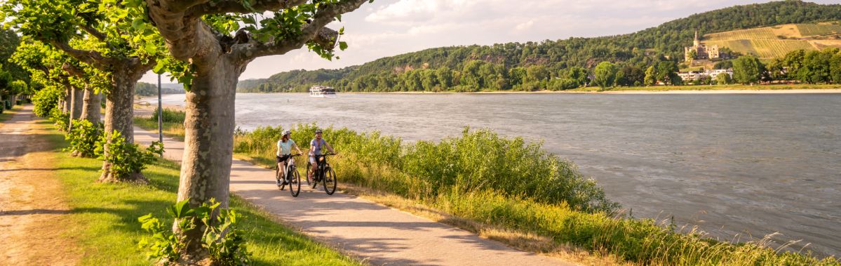 Ein grüner Weg entlang eines Flusses. Zwei Fahrradrfahrer sind auf dem Weg. Bäume befinden sich neben dem Weg und im Hintergrund ist eine grüne Landschaft zu erkennen.