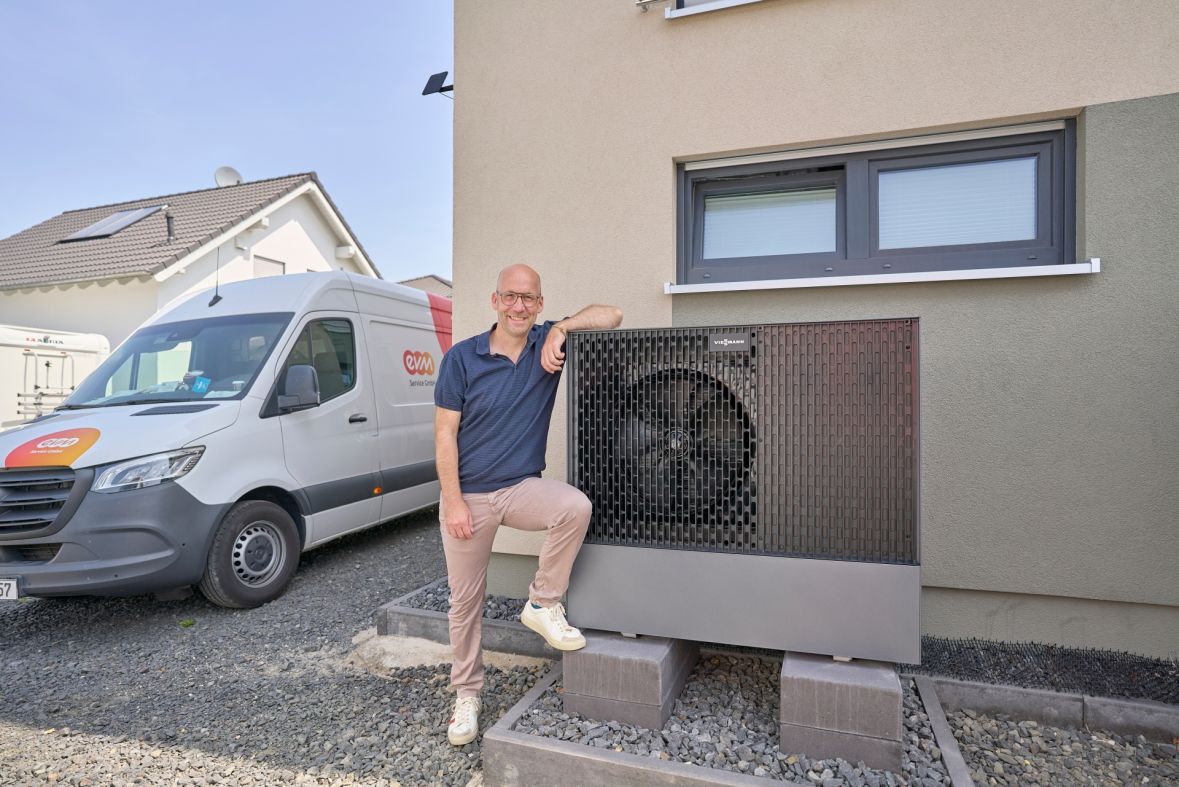 Thomas Schmitz steht neben einer außen installierten Wärmepumpe, die vor der Hauswand montiert ist. Im Hintergrund ist ein weißer Transporter mit „ewm“-Logo zu sehen.