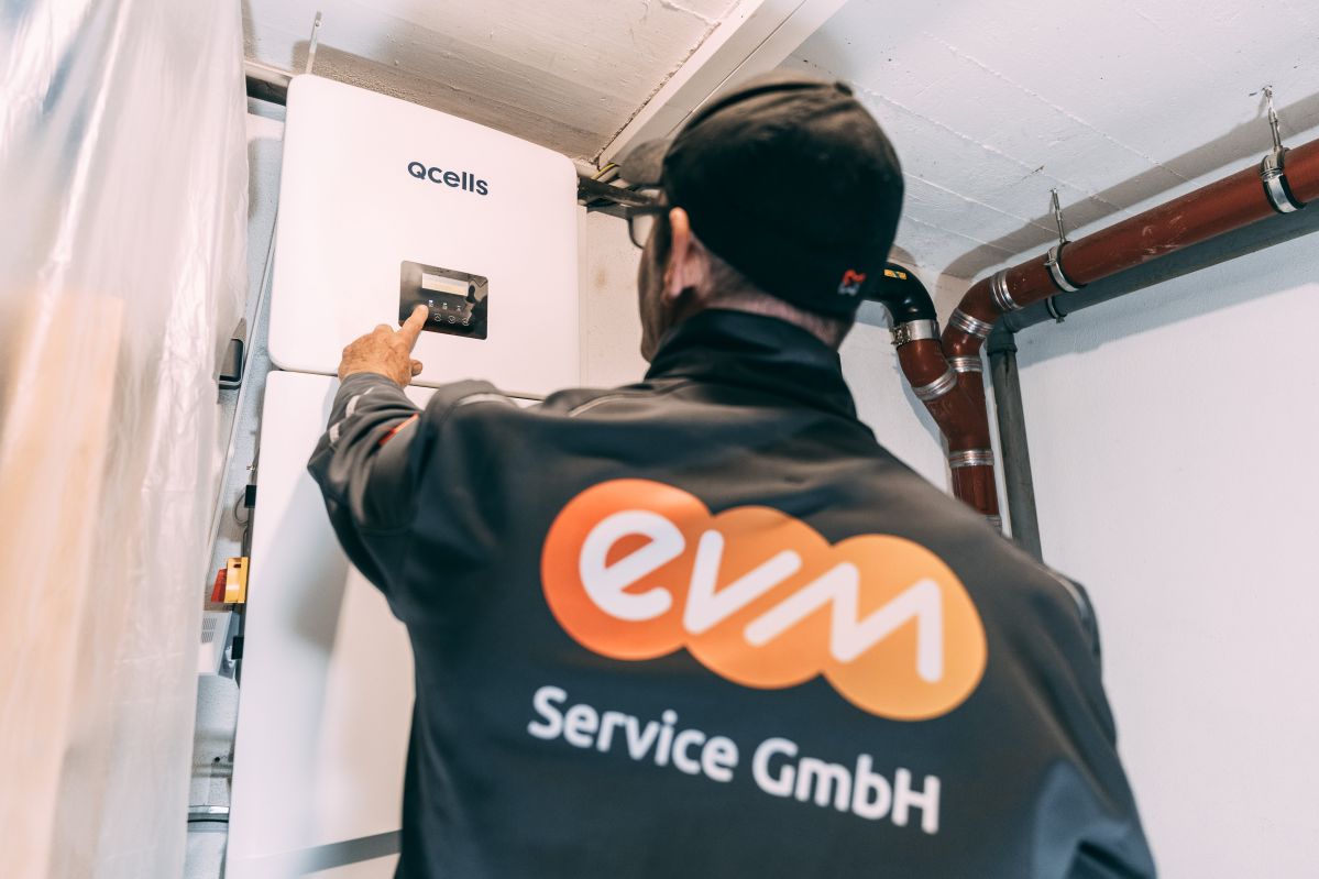 Einbau Wärmepumpe und PV Ein Mann mit einer grauen Jacke mit der Aufschrift "evm Service GmbH" arbeitet an einem Speicher in einem Heizungskeller.