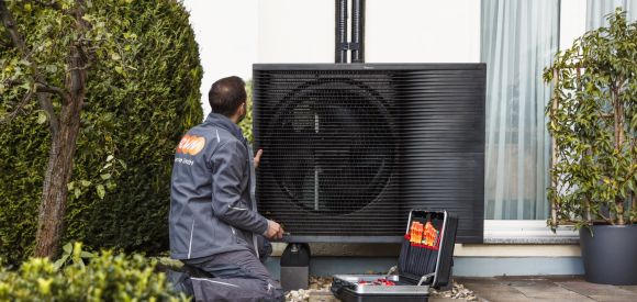 Ein Mann mit evm Service GmbH Arbeitskleidung kniet vor einer Wärmepumpe. Neben ihm steht ein Werkzeugkasten.