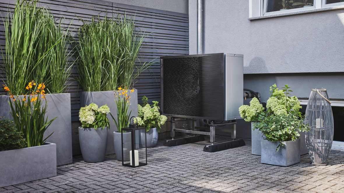 GEG: Was für Unternehmen relevant ist Eine modern aussehende Wärmepumpe steht auf einer gepflasterten Terrasse, umgeben von Blumentöpfen mit hohen Gräsern und blühenden Pflanzen neben einem Wohnhaus mit grauer Fassade.
