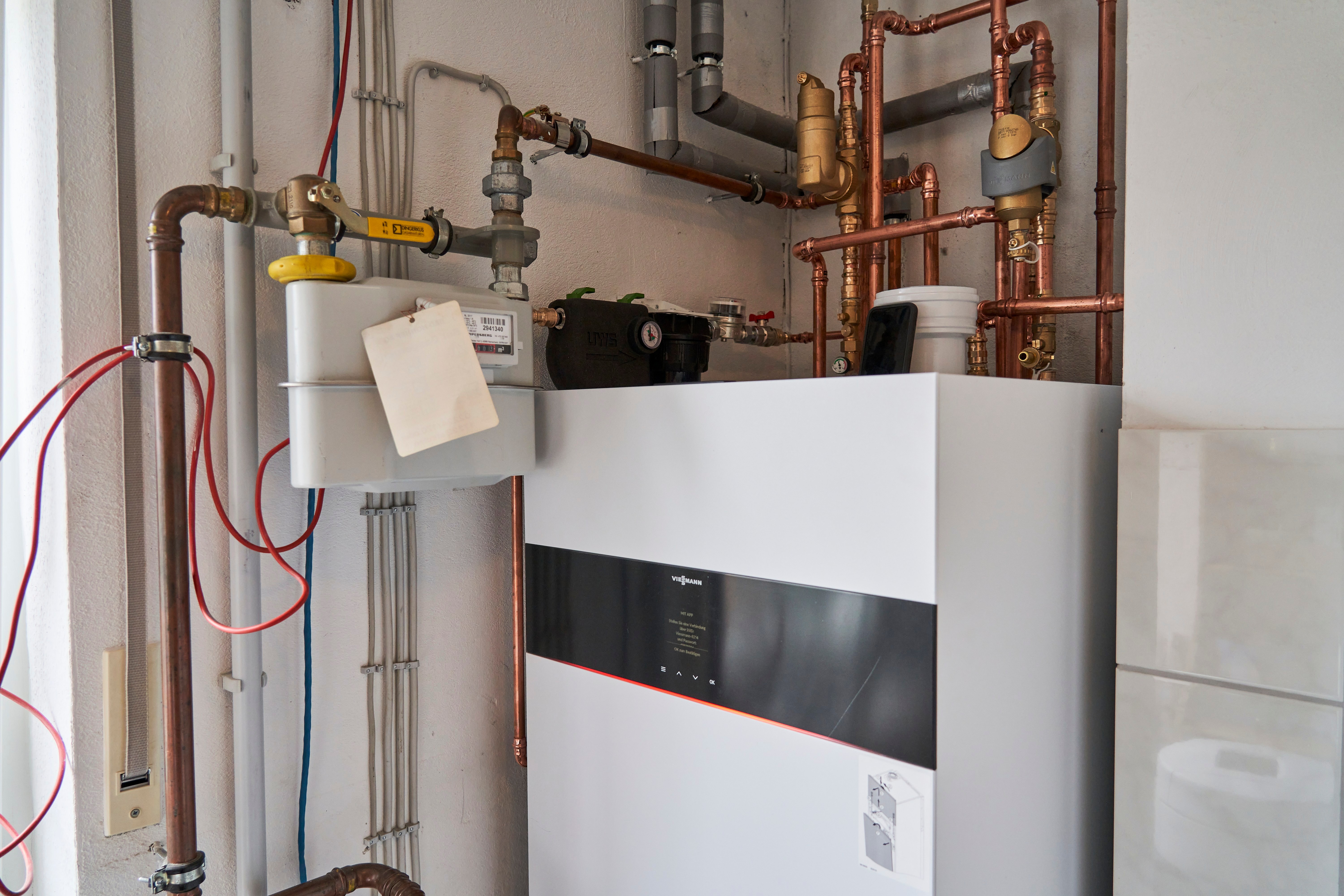 Unternehmen werden zum Energiesparen verpflichtet Ein moderner Heizkessel steht still in einem Technikraum, umgeben von zahlreichen Kupferrohren. Ein weißer Zettel hängt an einem Rohr, und Kabel laufen entlang der Wand.