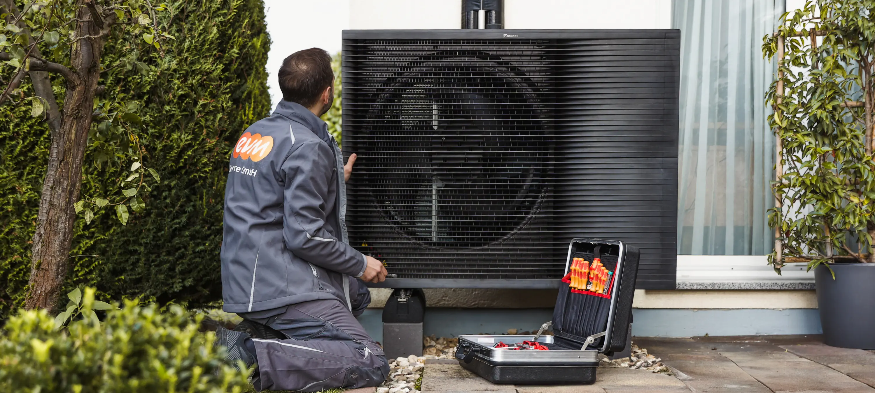 Ein Techniker überprüft eine große Wärmepumpe im Freien. Links im Bild stehen Pflanzen.