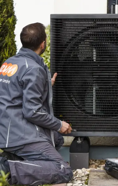 Ein Techniker überprüft eine große Wärmepumpe im Freien. Links im Bild stehen Pflanzen.