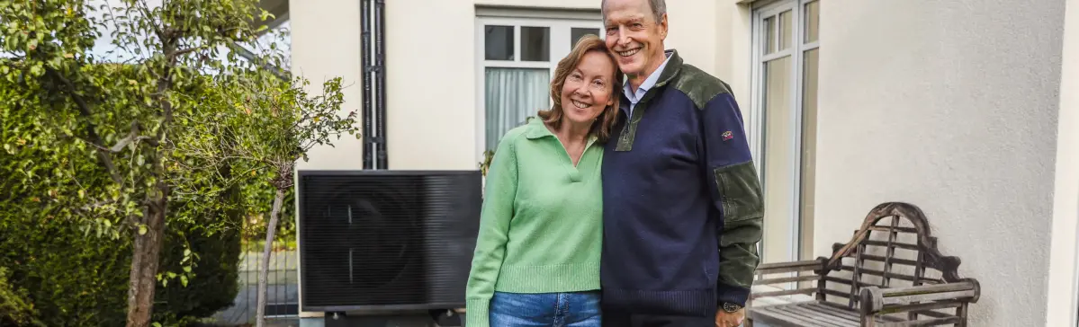 Ein älteres Paar steht vor einer Wärmepumpe in einem Garten und lächelt freundlich.