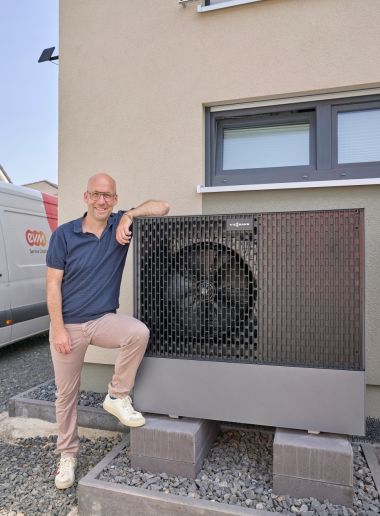 Wärmepumpe im Praxistest Thomas Schmitz steht neben einer außen installierten Wärmepumpe, die vor der Hauswand montiert ist. Im Hintergrund ist ein weißer Transporter mit „ewm“-Logo zu sehen.