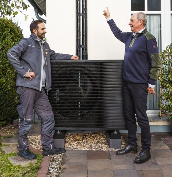 Ein Mann und ein evm Mitarbeiter stehen in einem Garten vor einer Wärmepumpe. Der Mann zeigt auf etwas während der Arbeiter an der Wärmepumpe lehnt.