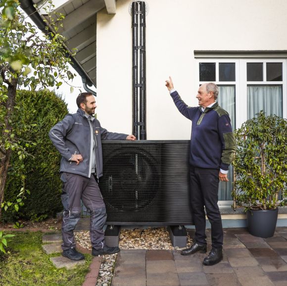 Ein Mann und ein evm Mitarbeiter stehen in einem Garten vor einer Wärmepumpe. Der Mann zeigt auf etwas während der Arbeiter an der Wärmepumpe lehnt.