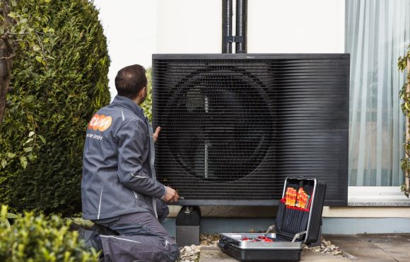 Ein Mann mit evm Service GmbH Arbeitskleidung kniet vor einer Wärmepumpe. Neben ihm steht ein Werkzeugkasten.