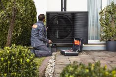 Ein Mann mit evm Service GmbH Arbeitskleidung kniet vor einer Wärmepumpe. Neben ihm steht ein Werkzeugkasten.