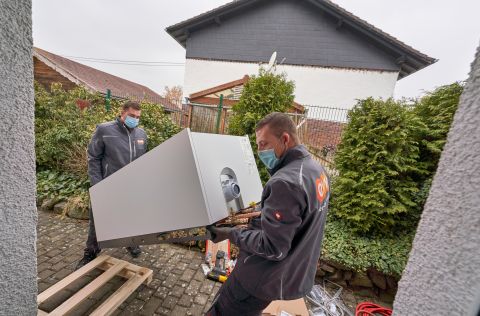 Trotz einiger Herausforderungen kann die neue Heizung problemlos eingebaut werden Zwei Monteure tragen die neue Heizanlage in das Haus des Kunden