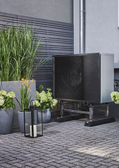 Eine modern aussehende Wärmepumpe steht auf einer gepflasterten Terrasse, umgeben von Blumentöpfen mit hohen Gräsern und blühenden Pflanzen neben einem Wohnhaus mit grauer Fassade.