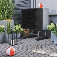 Eine Wärmepumpe steht in einem Garten vor einer Hauswand. Es befinden sich mehrere orangene Wasserbälle neben und  auf der Wärmepumpe. Es stehen moderne Pflanzen und eine Laterne daneben.