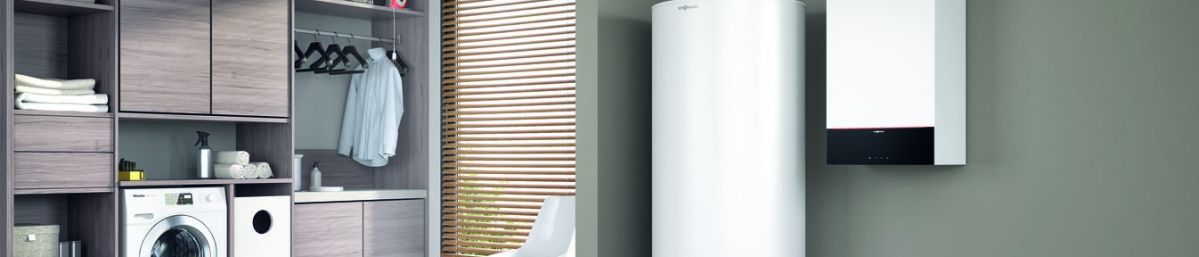 Eine große, weiße Heizung steht neben einem kleinen Wandgerät in einem modernen, minimalistisch eingerichteten Raum mit grüner Wand und Holzboden. Links ist ein Fenster mit Jalousie und ein Stuhl.
