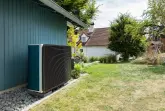 Eine Wärmepumpe steht an einer petrolfarbenen Hauswand in einem Garten mit einem grünen Rasen.