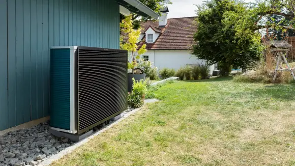 Eine Wärmepumpe steht an einer petrolfarbenen Hauswand in einem Garten mit einem grünen Rasen.