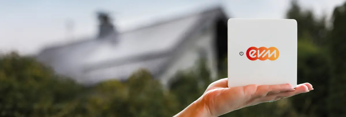 Eine Hand hält ein kleines weißes Gerät mit dem farbigen evm‑Logo vor einem unscharfen Hintergrund mit Bäumen und einem Hausdach.