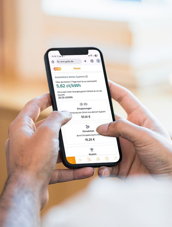 Eine Person hält ein Smartphone in beiden Händen und betrachtet in der evm‑App Energieinformationen wie Kosten, Einsparungen und Erträge auf einem hellen, unscharfen Hintergrund.