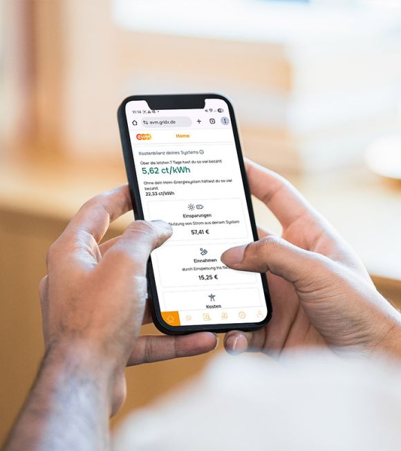 Eine Person hält ein Smartphone in beiden Händen und betrachtet in der evm‑App Energieinformationen wie Kosten, Einsparungen und Erträge auf einem hellen, unscharfen Hintergrund.