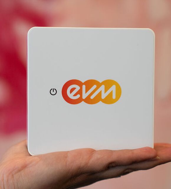 Eine Hand hält ein quadratisches, weißes Gerät mit dem farbigen „evm“-Logo