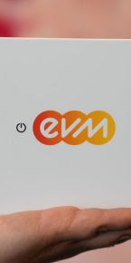 Eine Hand hält ein quadratisches, weißes Gerät mit dem farbigen „evm“-Logo