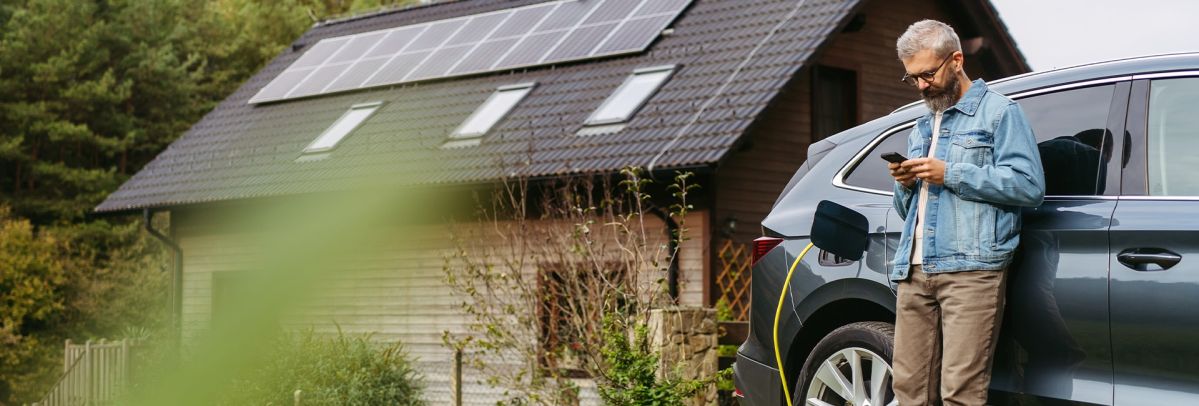 Eine Person lehnt an einem geparkten Elektroauto, das geladen wird, und schaut auf ein Smartphone. Im Hintergrund steht ein Haus mit großen Dachflächen, die vollständig mit Solarmodulen ausgestattet sind, umgeben von Bäumen.