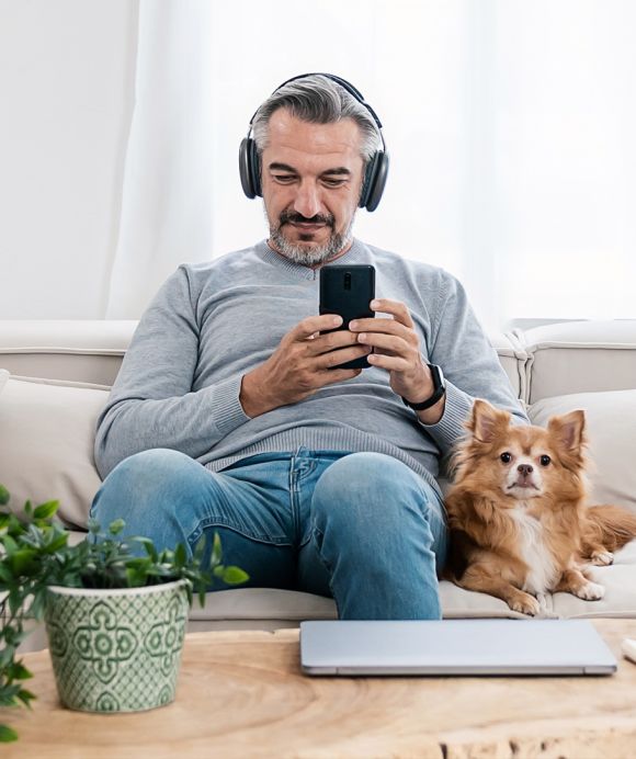 Eine Mann sitzt entspannt auf einem Sofa, trägt Kopfhörer und hält ein Smartphone in den Händen. Neben ihm liegt ein kleiner Hund, und auf dem Couchtisch stehen eine Pflanze und ein Laptop.