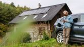 Klimafreundlich Im Grünen steht ein Haus mit einer Solaranlage, ein Mann steht an sein E-Auto gelehnt, das Auto wird geladen.