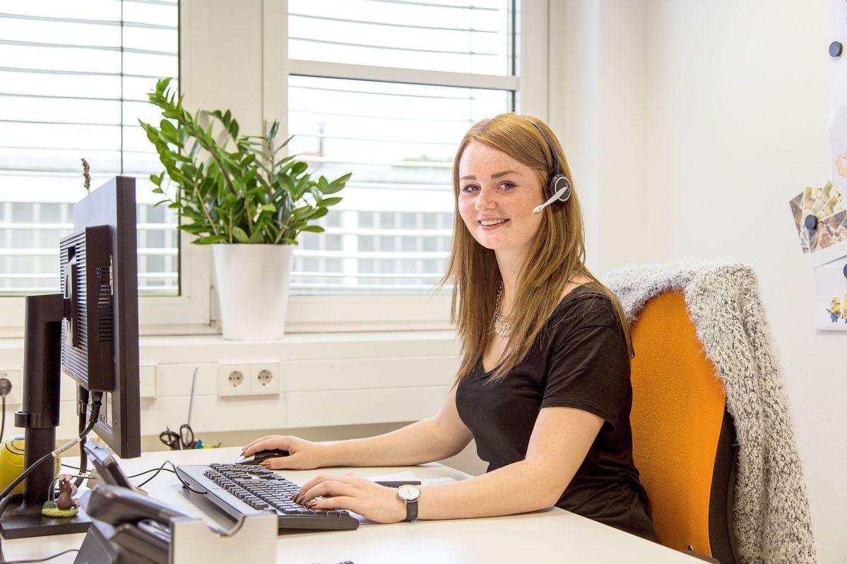 Eine Frau sitzt lächelnd an einem Schreibtisch und tippt auf einer Tastatur. Sie trägt ein Headset. Im Hintergrund stehen Fenster und eine Pflanze auf der Fensterbank.