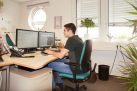 Eine Person sitzt am Schreibtisch, arbeitet an zwei Computermonitoren. Der Raum hat Fenster mit geschlossenen Jalousien, Pflanzen auf dem Fenstersims und einen runden Spiegel an der Wand.