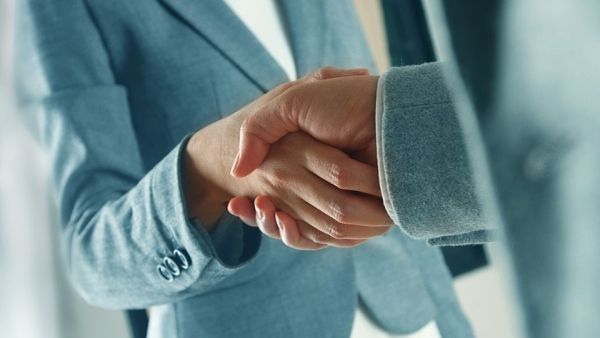 Nahaufnahme von zwei Personen die sich die Hand geben. Eine Person trägt einen blauen Blazer, die andere einen grauen Anzug.