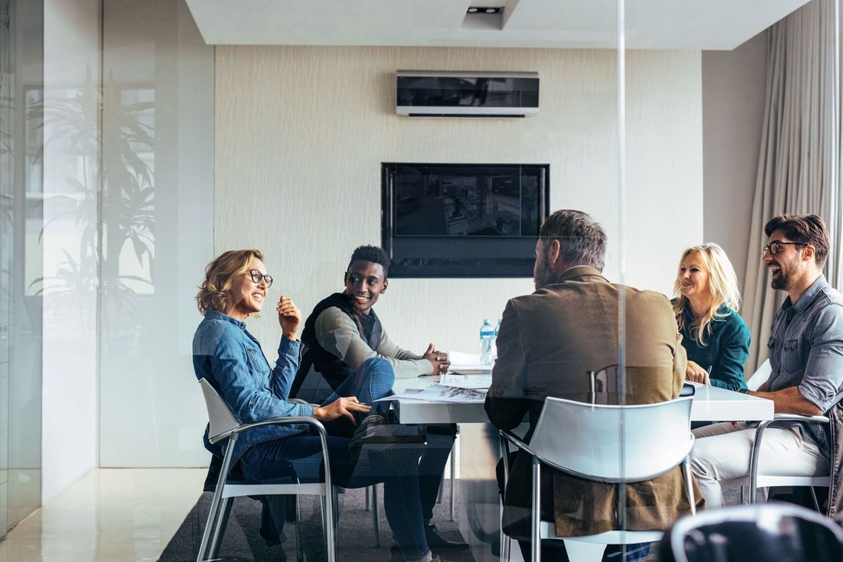 Konferenz im Büro Eine Gruppe von fünf Personen sitzt um einen Tisch und spricht miteinander in einem modernen, hellen Büro mit Glastüren und einem flachen Bildschirm an der Wand.