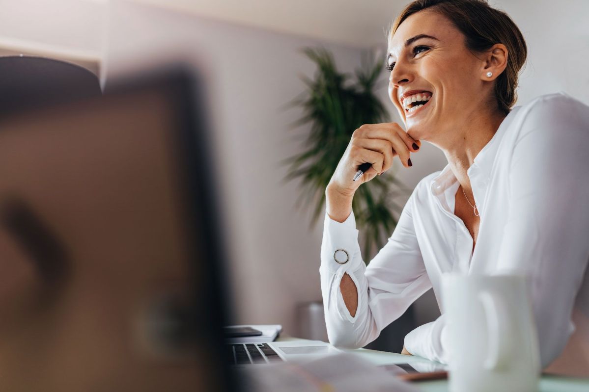 Frau im Büro lacht Eine Frau lächelt entspannt mit einem Stift in der Hand in einem modernen Büro, im Hintergrund ist eine grüne Pflanze verschwommen zu sehen.