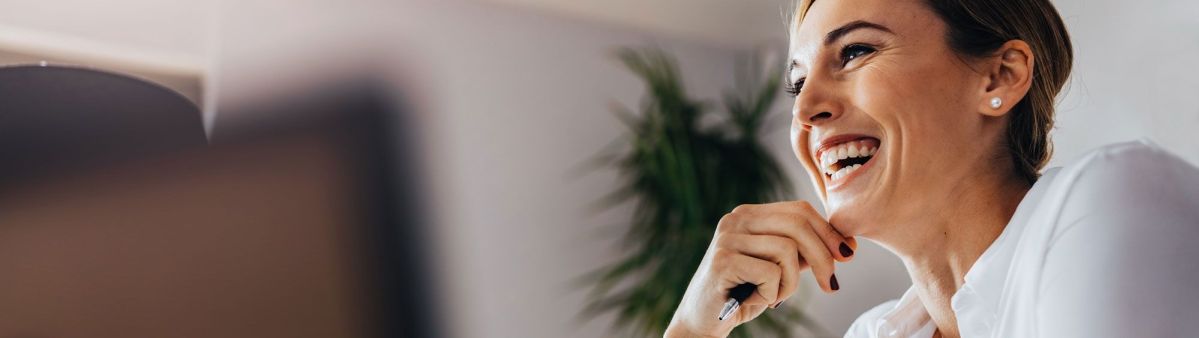 Eine Frau lächelt entspannt mit einem Stift in der Hand in einem modernen Büro, im Hintergrund ist eine grüne Pflanze verschwommen zu sehen.