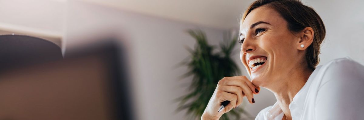 Eine Frau lächelt entspannt mit einem Stift in der Hand in einem modernen Büro, im Hintergrund ist eine grüne Pflanze verschwommen zu sehen.