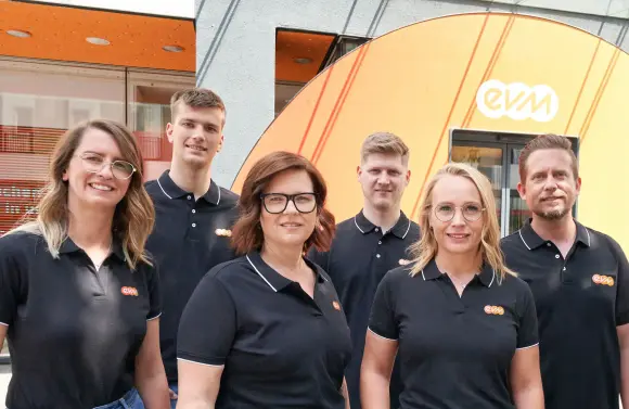 Drei Frauen und drei Männer tragen schwarze evm Poloshirts und lächeln freundlich. Sie stehen vor einem evm-Kundenzentrum.