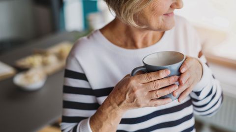 Eine ältere Frau hält eine graue Tasse in beiden Händen, lächelt leicht und trägt einen gestreiften Pullover. Im Hintergrund ist ein unscharfer Tisch zu sehen.
