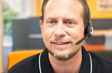 Ein Mann trägt ein Headset, lächelt und befindet sich in einem Büro mit orangefarbenen und grauen Elementen im Hintergrund.
