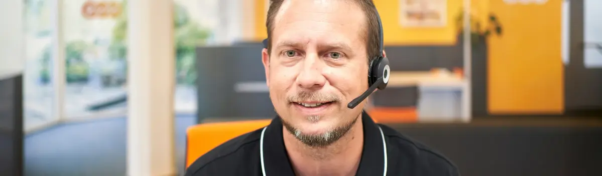 Ein Mann trägt ein Headset, lächelt und befindet sich in einem Büro mit orangefarbenen und grauen Elementen im Hintergrund.