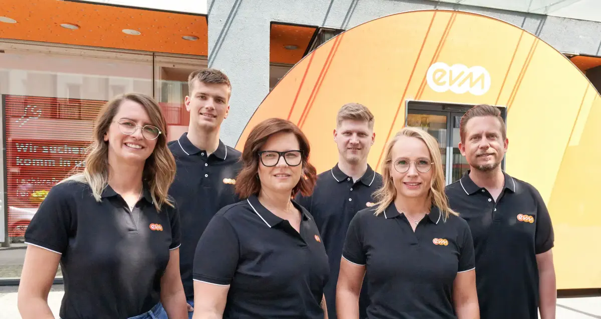 Drei Frauen und drei Männer tragen schwarze evm Poloshirts und lächeln freundlich. Sie stehen vor einem evm-Kundenzentrum.