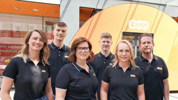 evm-KS Drei Frauen und drei Männer tragen schwarze evm Poloshirts und lächeln freundlich. Sie stehen vor einem evm-Kundenzentrum.