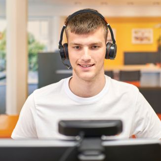 Ein Mann mit einem Headset sitzt vor einem Computer und lächelt freundlich. Er befindet sich in einem Büro.