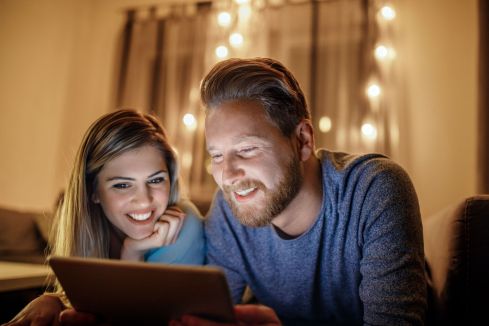 Eine Frau und ein Mann schauen lächelnd auf ei Tablet. Sie befinden sich in einem Wohnzimmer. Im Hintergrund ist eine Lichterkette zu erkennen.