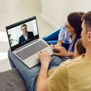 Ein Laptop zeigt eine Videokonferenz mit einem lächelnden Mann im Anzug. Eine Familie sitzt nah beisammen auf einem Sofa und schaut konzentriert auf den Bildschirm.