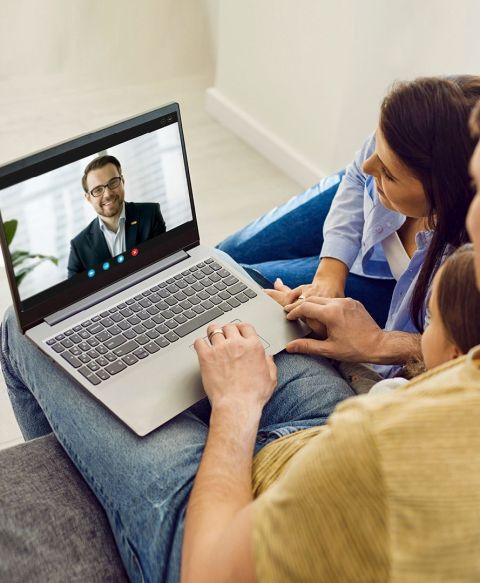 Ein Laptop zeigt eine Videokonferenz mit einem lächelnden Mann im Anzug. Eine Familie sitzt nah beisammen auf einem Sofa und schaut konzentriert auf den Bildschirm.