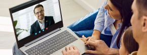 Ein Laptop zeigt eine Videokonferenz mit einem lächelnden Mann im Anzug. Eine Familie sitzt nah beisammen auf einem Sofa und schaut konzentriert auf den Bildschirm.
